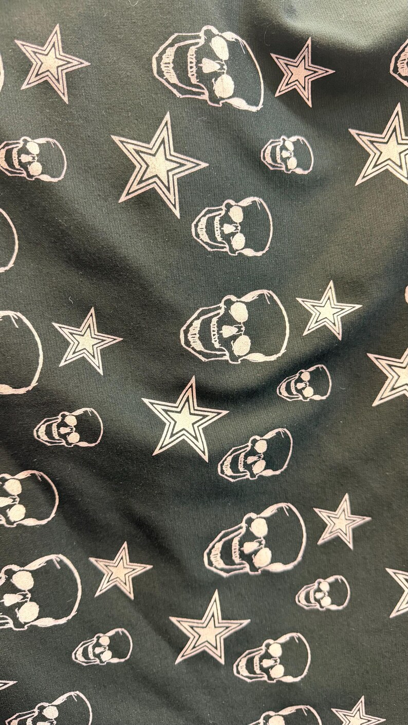 May include: Dark green fabric featuring a repeating pattern of white skulls and five-pointed stars. The skulls have eye sockets and teeth, and the stars have a double outline.