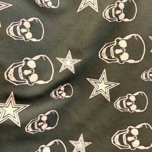 May include: Dark grey fabric featuring a repeating pattern of white skulls and five-pointed stars. The skulls are detailed, and the stars have a classic outline. The design is evenly spaced across the material, creating a striking visual.