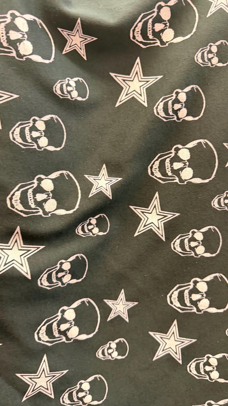 May include: Dark grey fabric featuring a repeating pattern of white skulls and five-pointed stars. The skulls are detailed, and the stars have a classic outline. The design is evenly spaced across the material, creating a striking visual.