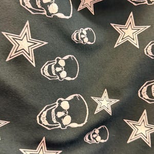 May include: Dark fabric featuring a repeating pattern of white skulls and stars. The skulls have eye sockets, a nose cavity, and teeth. The stars have a five-point design.