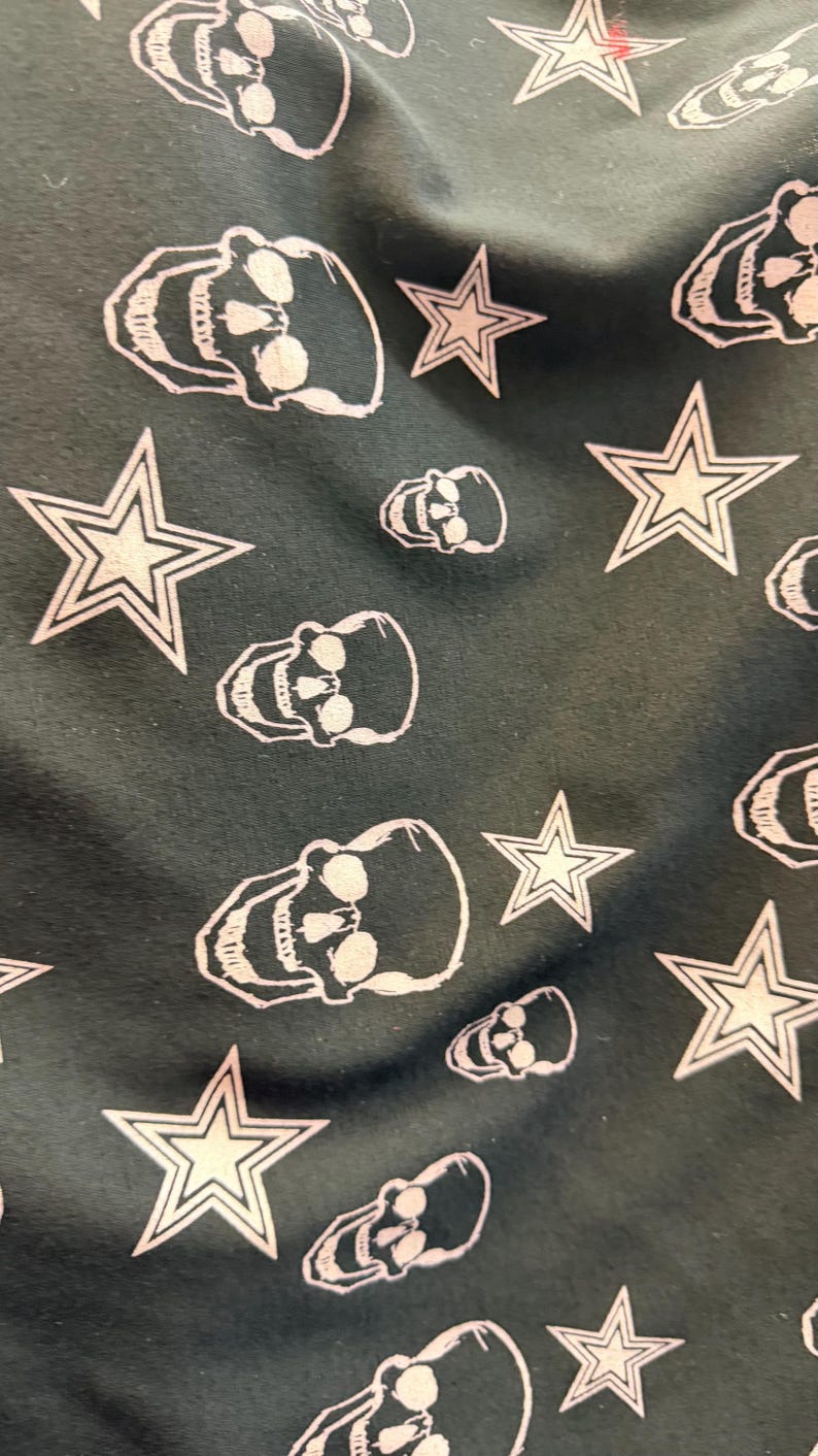 May include: Dark fabric featuring a repeating pattern of white skulls and stars. The skulls have eye sockets, a nose cavity, and teeth. The stars have a five-point design.