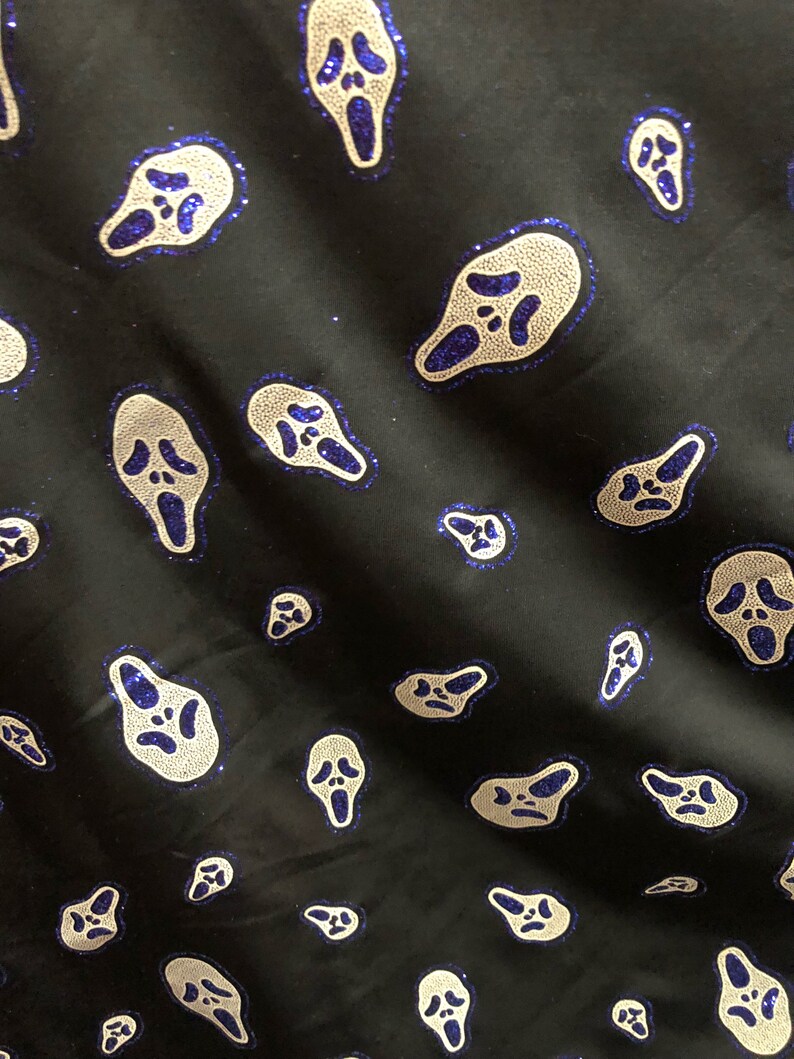 May include: Black fabric with a repeating pattern of white and blue glitter ghost faces. The ghost faces are in a scream pose with their mouths open.