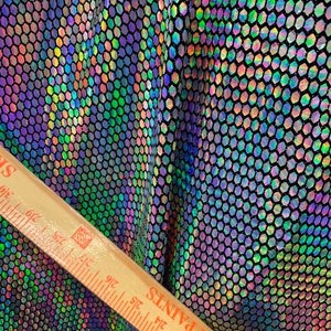 May include: Black fabric with a holographic hexagonal pattern. The fabric is iridescent and shimmers in different colors, including green, blue, purple, and pink. A wooden ruler is placed on the fabric to show the scale.