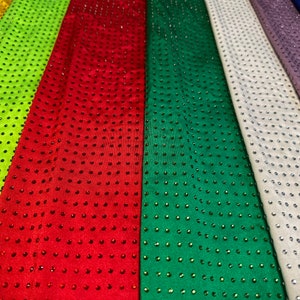 Op de afbeelding: Meerdere stroken stof naast elkaar in geel, limoengroen, rood, groen, wit, lavendel en blauw. Elke strook is voorzien van een uniform raster van kleine, reflecterende strass-steentjes die overeenkomen met de kleur van de stof.