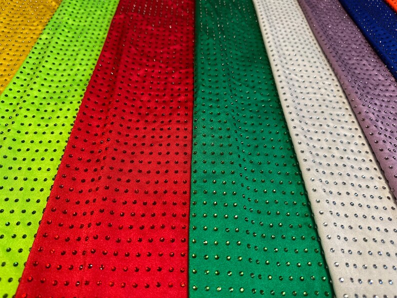 Op de afbeelding: Meerdere stroken stof naast elkaar in geel, limoengroen, rood, groen, wit, lavendel en blauw. Elke strook is voorzien van een uniform raster van kleine, reflecterende strass-steentjes die overeenkomen met de kleur van de stof.
