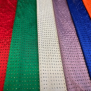 Op de afbeelding: Een verzameling verticaal gerangschikte stroken stof in rood, groen, wit, lavendel, blauw en oranje. Elke strook is voorzien van een uniform raster van kleine, reflecterende, ronde versieringen die een consistent stippenpatroon vormen.