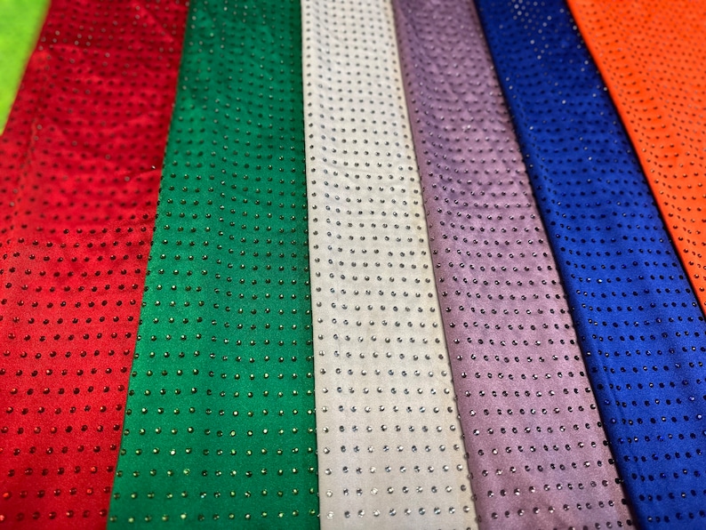 Op de afbeelding: Een verzameling verticaal gerangschikte stroken stof in rood, groen, wit, lavendel, blauw en oranje. Elke strook is voorzien van een uniform raster van kleine, reflecterende, ronde versieringen die een consistent stippenpatroon vormen.