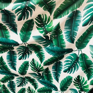 Op de afbeelding: Een witte stof met een herhalend patroon van groene tropische bladeren. De bladeren zijn in verschillende vormen en maten, waaronder palmbladeren en monstera bladeren.