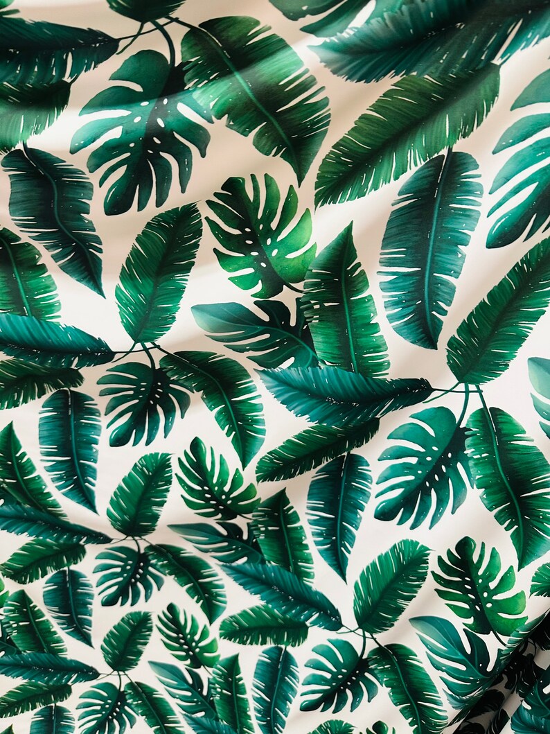 Op de afbeelding: Een witte stof met een herhalend patroon van groene tropische bladeren. De bladeren zijn in verschillende vormen en maten, waaronder palmbladeren en monstera bladeren.