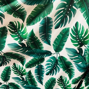 Op de afbeelding: Witte stof met een herhalend patroon van groene tropische bladeren. De bladeren zijn in verschillende vormen en maten, waaronder grote monstera bladeren en kleinere palmbladeren.
