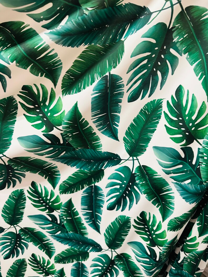 Op de afbeelding: Witte stof met een herhalend patroon van groene tropische bladeren. De bladeren zijn in verschillende vormen en maten, waaronder grote monstera bladeren en kleinere palmbladeren.