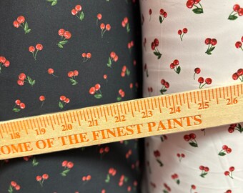 Puede incluir: Dos rollos de tela con estampado de cerezas. La tela de la izquierda es negra con cerezas rojas. La tela de la derecha es blanca con cerezas rojas. Una regla de madera con el texto "Home of the Finest Paints" está colocada sobre los rollos de tela.