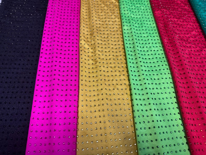 Op de afbeelding: Zes verticale stroken stof in zwart, felroze, goud, limoengroen, rood en donkerblauwgroen. Elke strook is voorzien van een uniform raster van kleine, reflecterende, ronde studs of strass-steentjes die een consistent, herhalend patroon vormen.