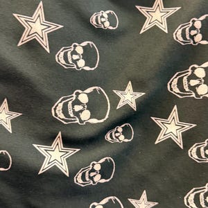May include: Dark green fabric featuring a repeating pattern of white skulls and five-pointed stars. The skulls have eye sockets, teeth, and a jawline. The stars have a white outline and a white centre.