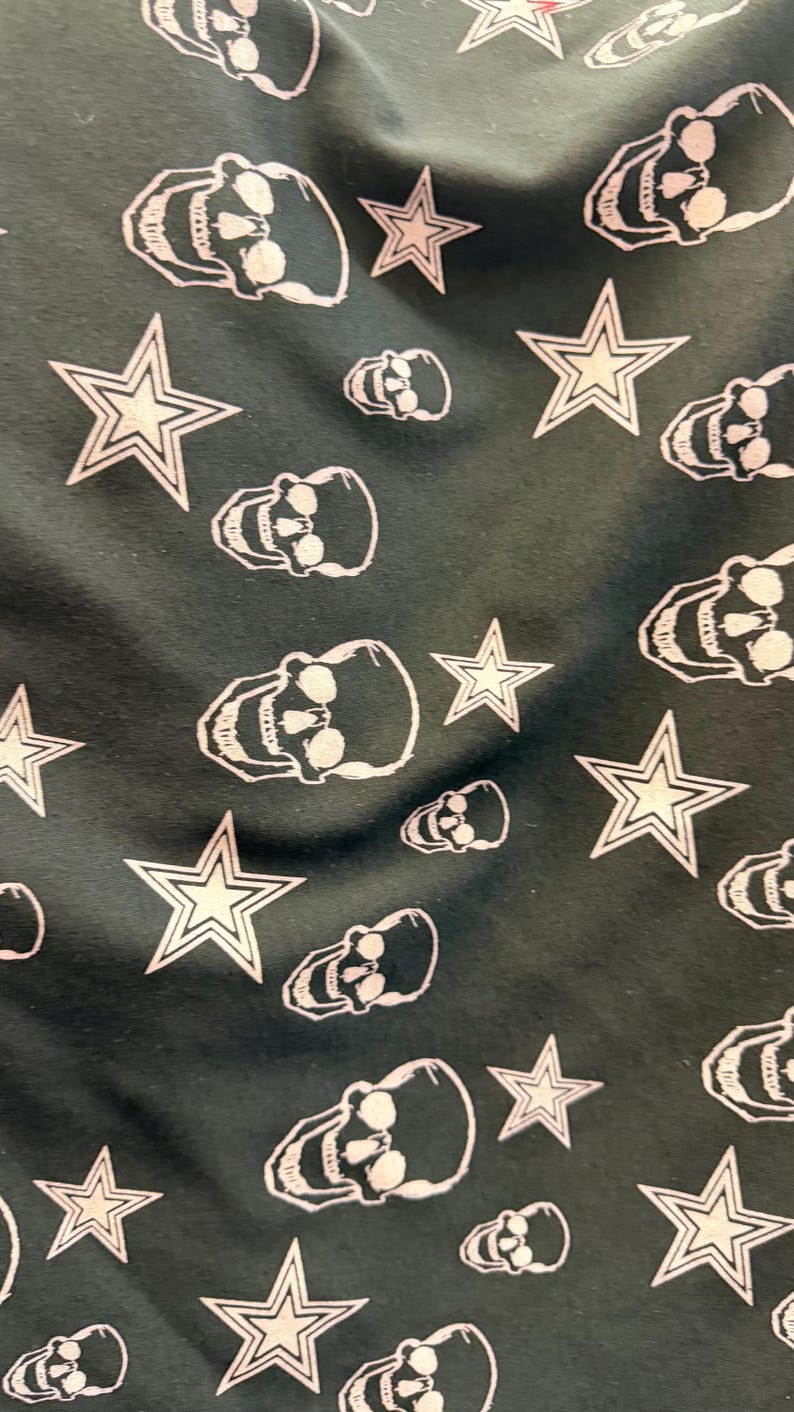 May include: Dark green fabric featuring a repeating pattern of white skulls and five-pointed stars. The skulls have eye sockets, teeth, and a jawline. The stars have a white outline and a white centre.