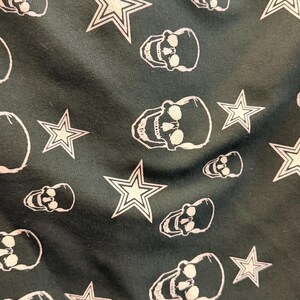 May include: Dark green fabric featuring a repeating pattern of white skulls and stars. The skulls are detailed, and the stars have a five-point design. The design has a gothic or edgy theme.