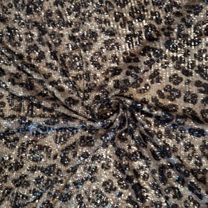May include: Close-up of a fabric with a leopard print pattern. The fabric has a tan background with black spots. The fabric is shiny and has a textured appearance. The fabric is likely used for clothing or upholstery.