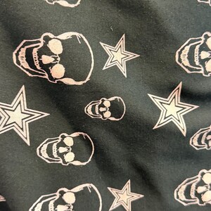 May include: Dark fabric featuring a repeating pattern of white skulls and stars. The skulls have detailed facial features, and the stars have a five-point design. The pattern is evenly spaced across the surface.