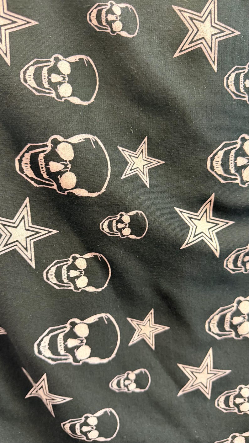 May include: Dark fabric featuring a repeating pattern of white skulls and stars. The skulls have detailed facial features, and the stars have a five-point design. The pattern is evenly spaced across the surface.