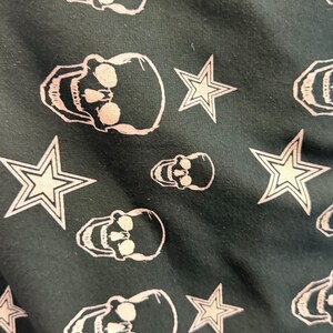 May include: Dark green fabric featuring a repeating pattern of white skulls and star shapes. The skulls are detailed with eye sockets and teeth, and the stars have a five-point design. The fabric has a slightly textured appearance.