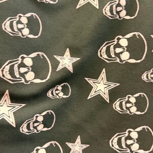 May include: Dark green fabric featuring a repeating pattern of white skulls and stars. The skulls are detailed with eye sockets and teeth, and the stars have a five-point design. The design has a gothic and Americana aesthetic.