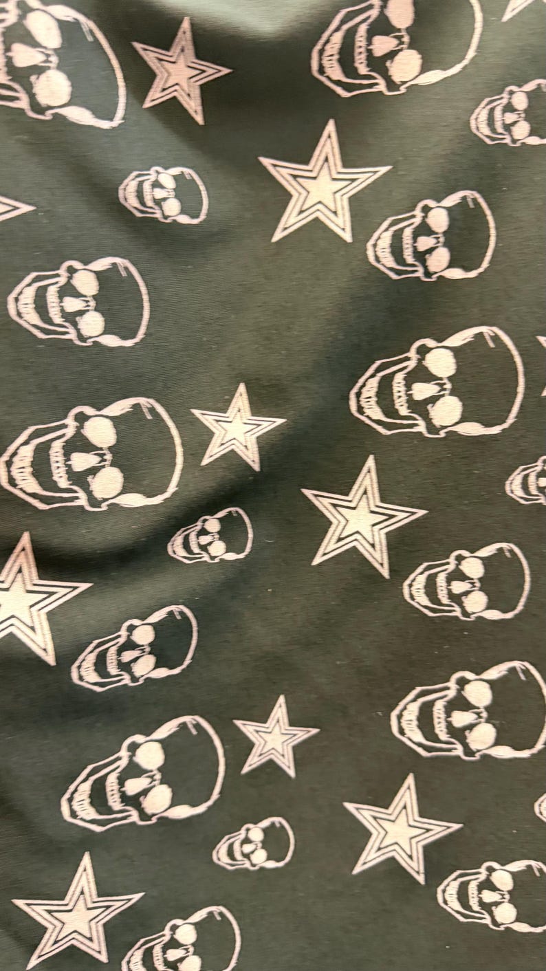 May include: Dark green fabric featuring a repeating pattern of white skulls and stars. The skulls are detailed with eye sockets and teeth, and the stars have a five-point design. The design has a gothic and Americana aesthetic.