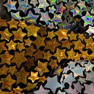 May include: A black fabric background with a variety of gold and iridescent star-shaped patches. The stars are sewn on the fabric and have a glittery finish.