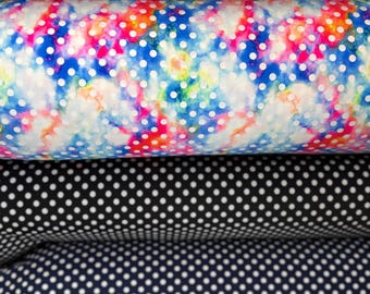 May include: A stack of fabric rolls with various polka dot patterns. The top roll features white fabric with black dots. Below it is a roll with a colorful tie-dye pattern. Other rolls include black and navy fabrics with white dots, and a neon pink and green pattern.
