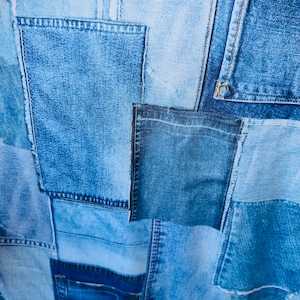 Op de afbeelding: Een patchwork stof gemaakt van verschillende tinten blauwe denim. De stof is gemaakt van vierkanten en rechthoeken van denim die aan elkaar zijn genaaid.
