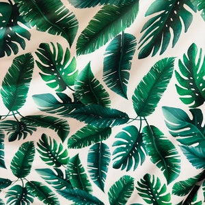 Op de afbeelding: Witte stof met een herhalend patroon van groene tropische bladeren. De bladeren zijn in verschillende vormen en maten, waaronder grote monstera bladeren en kleinere palmbladeren.