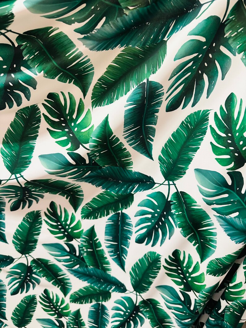 Op de afbeelding: Witte stof met een herhalend patroon van groene tropische bladeren. De bladeren zijn in verschillende vormen en maten, waaronder grote monstera bladeren en kleinere palmbladeren.