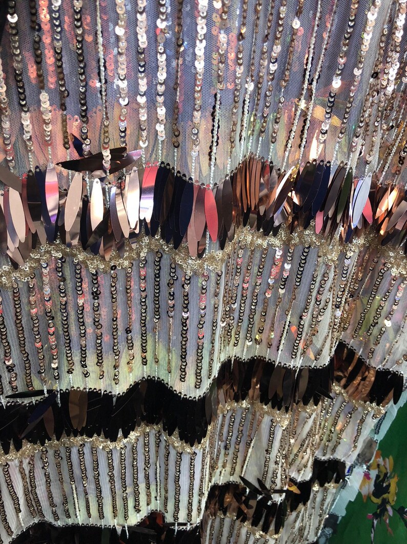 K&ouml;nnte beinhalten: Nahaufnahme eines Stoffes mit einem mehrschichtigen Design aus Pailletten und metallischen Bl&auml;ttern. Der Stoff ist ein durchsichtiges Netz mit wei&szlig;em Hintergrund. Die Pailletten sind in Gold-, Silber- und Schwarzt&ouml;nen gehalten. Die metallischen Bl&auml;tter sind in Kupfer- und Goldt&ouml;nen gehalten.