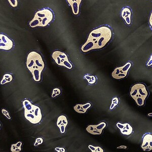 May include: Black fabric with a repeating pattern of white ghost faces with blue outlines. The ghost faces are all the same size and shape, and they are evenly spaced apart.