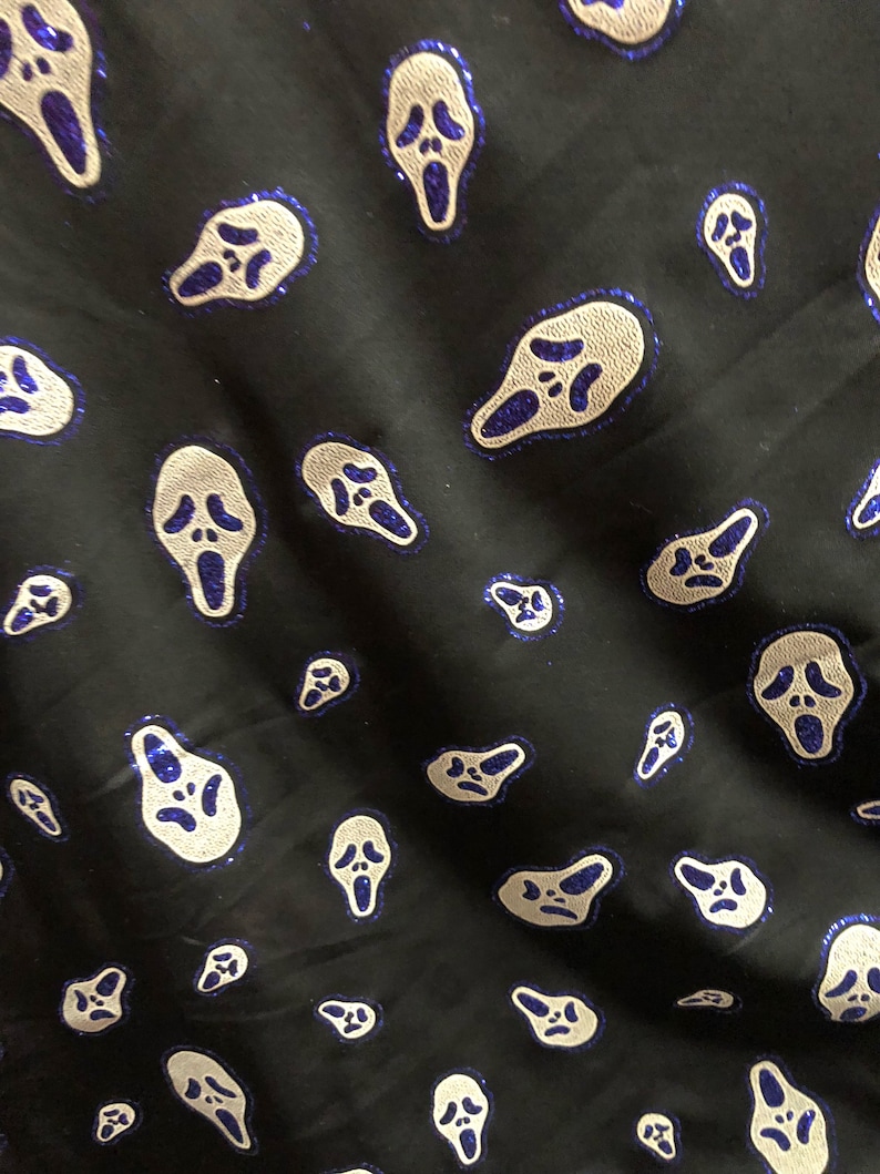 May include: Black fabric with a repeating pattern of white ghost faces with blue outlines. The ghost faces are all the same size and shape, and they are evenly spaced apart.