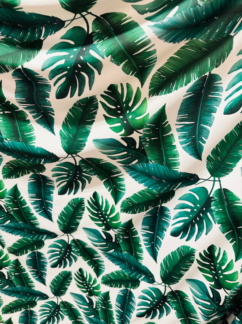 Op de afbeelding: Een witte stof met een herhalend patroon van groene tropische bladeren. De bladeren zijn in verschillende vormen en maten, waaronder grote monstera bladeren en kleinere palmbladeren.