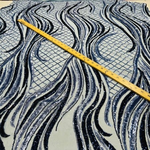 Puede incluir: Una tela con un patrón de ondas en tonos azules y negros, adornada con lentejuelas. El diseño incluye un patrón similar a una cuadrícula. Una regla de madera se coloca en diagonal sobre la tela.