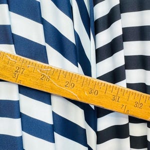 K&ouml;nnte beinhalten: Nahaufnahme von gestreiftem Stoff in Marineblau und Schwarz mit wei&szlig;en Streifen. Ein Holzlineal zeigt Messungen von 68,5 cm bis 81,3 cm. Der Stoff ist drapiert und zeigt Textur und Muster.