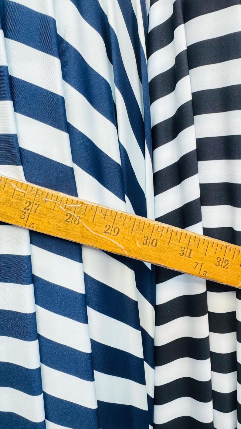 K&ouml;nnte beinhalten: Nahaufnahme von gestreiftem Stoff in Marineblau und Schwarz mit wei&szlig;en Streifen. Ein Holzlineal zeigt Messungen von 68,5 cm bis 81,3 cm. Der Stoff ist drapiert und zeigt Textur und Muster.