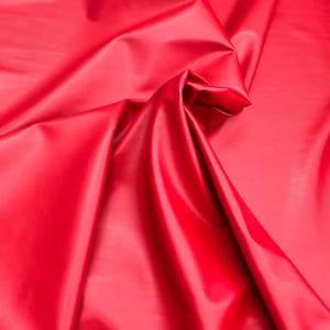 Può includere: Primo piano di un tessuto liscio rosso con una texture morbida e setosa. Il tessuto è piegato e drappeggiato, creando un aspetto lussuoso ed elegante.