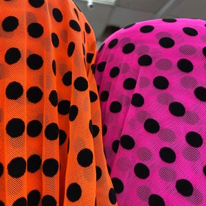 May include: Close-up of two fabrics: one bright orange, the other hot pink. Both are sheer and patterned with large, evenly spaced black polka dots. The fabric is draped, creating folds and shadows that highlight the texture and color contrast.