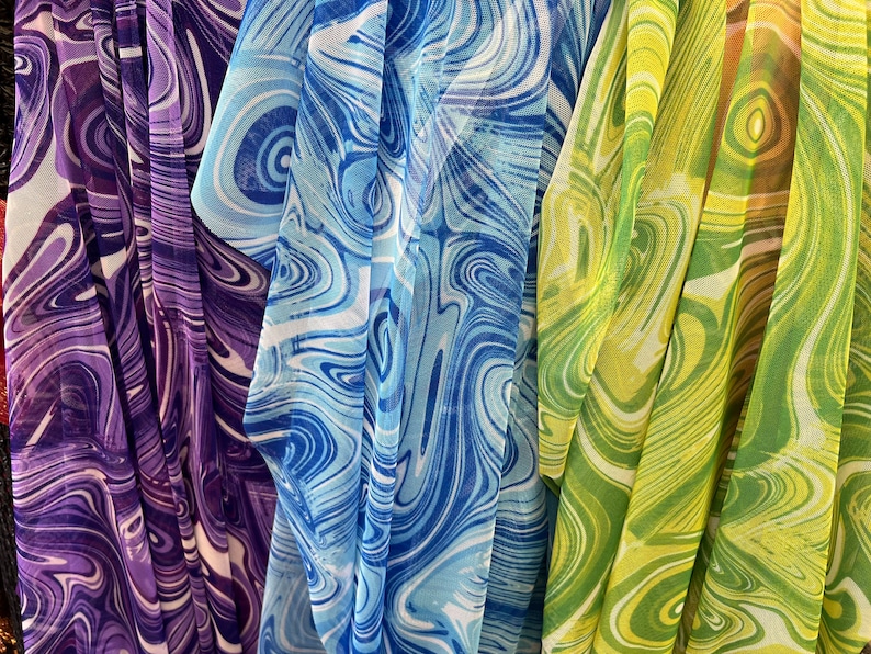 K&ouml;nnte beinhalten: Nahaufnahme eines farbenfrohen Stoffes mit einem Wirbelmuster. Der Stoff ist in vertikalen Falten drapiert und zeigt Schattierungen von Lila, Blau und Gr&uuml;n. Das Design &auml;hnelt einem Marmoreffekt und erzeugt eine dynamische und flie&szlig;ende visuelle Textur. Der Stoff wirkt leicht und transparent.
