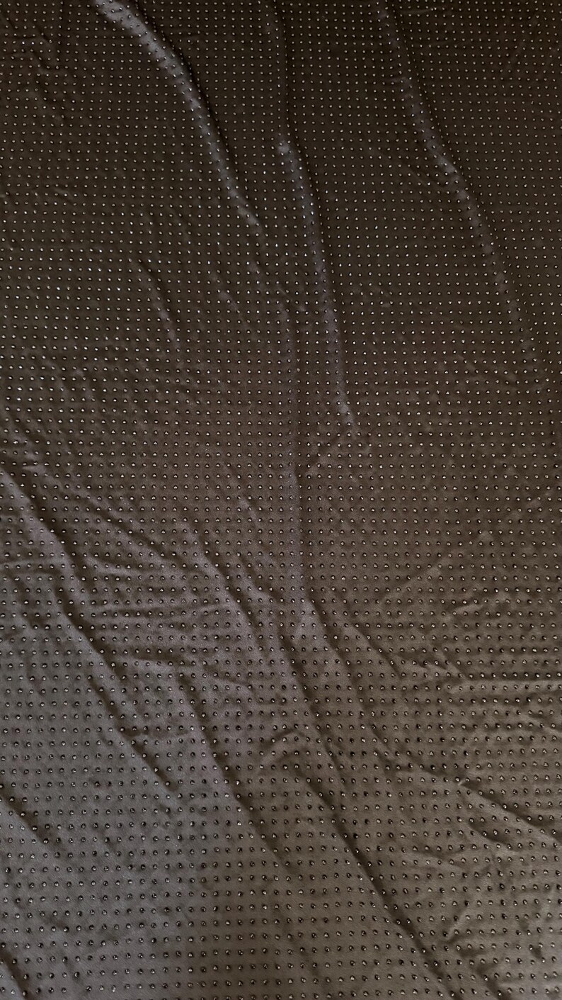 Op de afbeelding: Een donkerbruine stof met een uniform raster van kleine, verhoogde, cirkelvormige stippen over het gehele oppervlak. Het textiel vertoont subtiele vouwen en kreukels, wat zorgt voor een getextureerd uiterlijk met een consistent patroon.