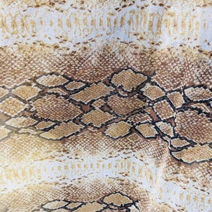 Könnte beinhalten: Nahaufnahme eines Stoffes mit Schlangenmuster. Das Muster zeigt ein sich wiederholendes Design aus braunen, beigen und cremefarbenen Schuppen. Der Stoff wirkt leicht und fließend, mit leichtem Glanz.