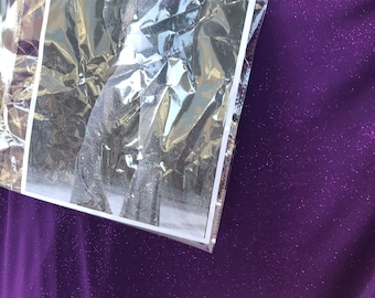 May include: A close-up of a purple fabric with a sparkly texture. A clear plastic bag with a black and white photo of a person singing into a microphone is on top of the fabric.