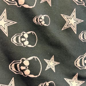 May include: Dark teal fabric featuring a repeating pattern of white skulls and five-pointed stars. The skulls have eye sockets, teeth, and a rounded cranium. The stars have a white outline and a filled-in centre.