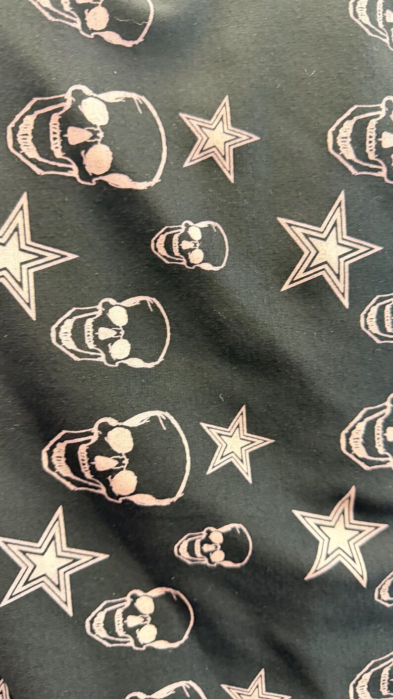May include: Dark teal fabric featuring a repeating pattern of white skulls and five-pointed stars. The skulls have eye sockets, teeth, and a rounded cranium. The stars have a white outline and a filled-in centre.