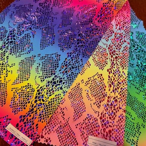 Könnte beinhalten: Nahaufnahme mehrerer Stoffmuster mit Schlangenmuster. Die Stoffe zeigen einen lebendigen Farbverlauf, darunter Lila, Rosa, Gelb und Grün. Die Muster haben eine glänzende, schillernde Oberfläche.