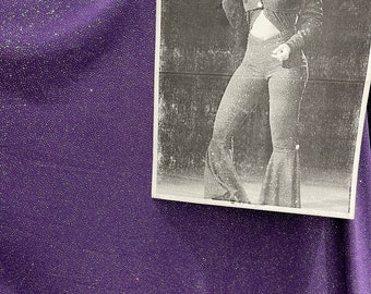 May include: A black and white photo of a person singing into a microphone. The person is wearing a sparkly top and pants. The photo is taped to a purple sparkly fabric.