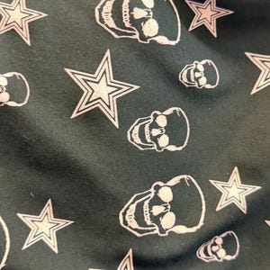 May include: Dark green fabric featuring a repeating pattern of white skulls and stars. The skulls are detailed, and the stars have a five-point design. The fabric has a gothic aesthetic, suitable for clothing or accessories.