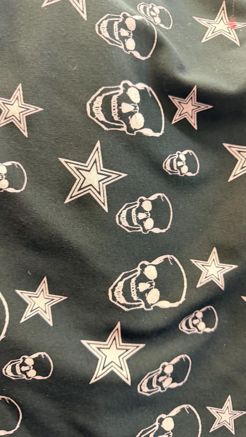 May include: Dark green fabric featuring a repeating pattern of white skulls and stars. The skulls are detailed, and the stars have a five-point design. The fabric has a gothic aesthetic, suitable for clothing or accessories.